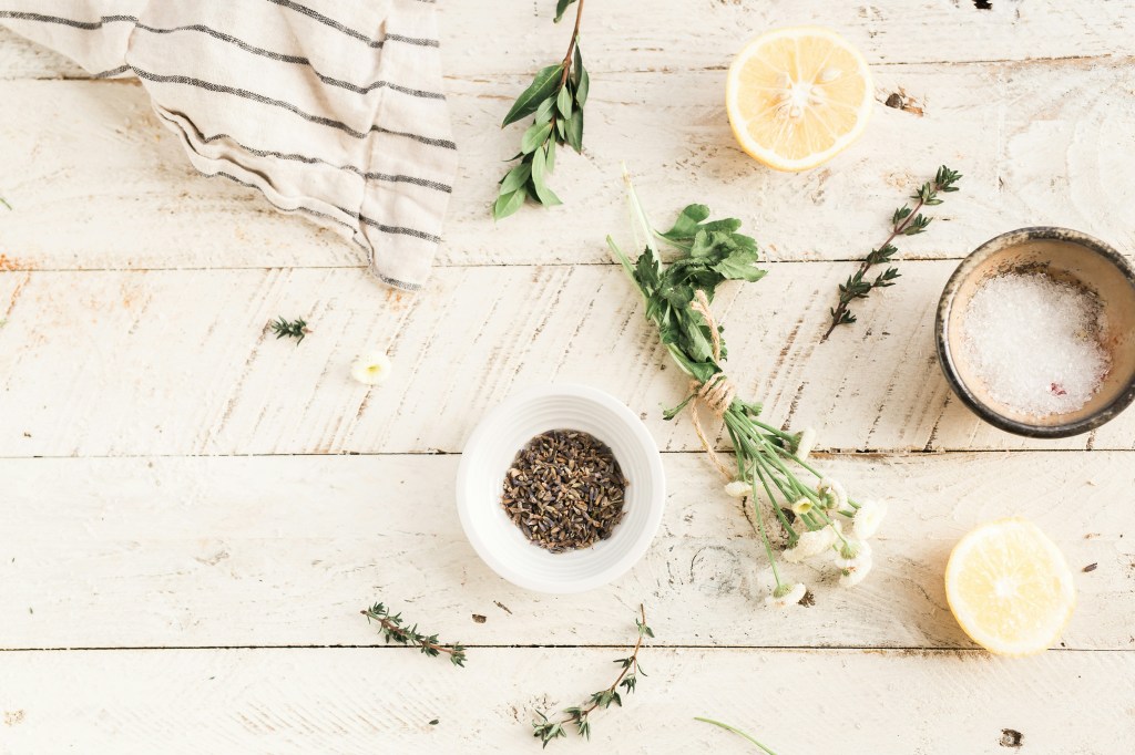 A wooden countertop with various ingredients including a bowl of lavender, fresh herbs, lemon halves, and a small bowl of salt, accompanied by a striped kitchen towel.
